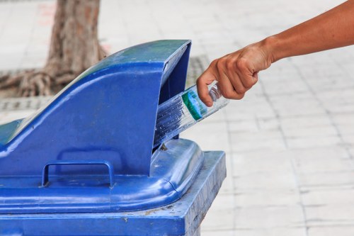 A person's hand is seen holding a plastic bottle and placing it into a blue recycling bin, which has its lid partially open. The bin is situated on a paved pavement area, with a tree trunk visible in the background, indicating an outdoor urban or suburban environment. The hand appears to belong to an adult, and the bottle is clear with a green label. The image highlights proper waste disposal practices, emphasizing recycling, and is relevant to rubbish removal services offered by companies like Garden Clearance Fulham, based near Fulham. The scene is well-lit, suggesting daytime, and the focus is on the interaction between the person and the recycling container, with the surrounding pavement and natural setting providing contextual environment for household or community waste management within the Fulham area.
