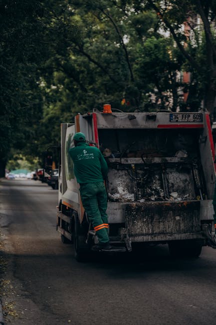 A waste collection worker in a green uniform and safety helmet stands at the rear of a commercial rubbish collection truck on a residential street, preparing for waste loading. The truck features a la
