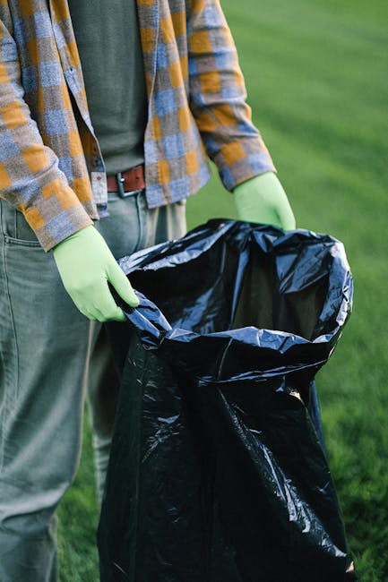 A waste collection worker in a green uniform and safety helmet stands at the rear of a commercial rubbish collection truck on a residential street, preparing for waste loading. The truck features a la