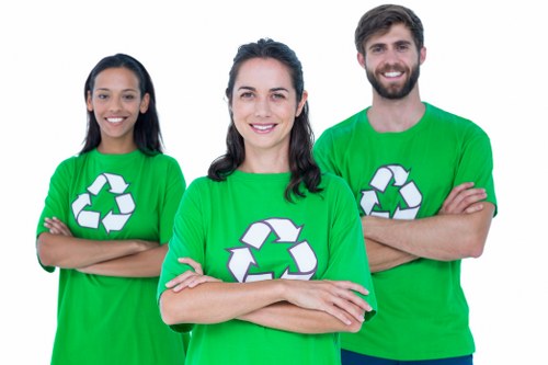 A team of three waste management professionals wearing bright green T-shirts featuring a white recycling symbol are standing outdoors on a paved surface. The woman in the center has shoulder-length dark hair and is smiling with arms crossed. To her left, a woman with long dark hair also crosses her arms and smiles warmly. To her right, a man with short dark hair and a beard has his arms crossed and is smiling. The background suggests a garden or outdoor area, possibly part of a garden clearance service in Fulham, with natural light illuminating the scene. Their attire and confident posture indicate they are prepared for rubbish removal or clearance tasks, aligning with services provided by Garden Clearance Fulham in the Fulham area, known for professional waste disposal solutions in London.